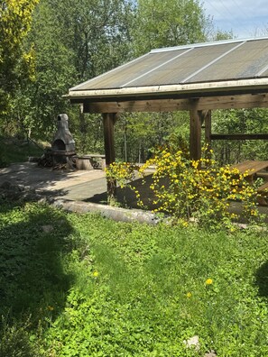 A rustic outdoor area with a wooden structure, a stone oven, and a garden with yellow flowers. - Charming 3-room apartment in the hills above Gérardmer (Gérardmer)