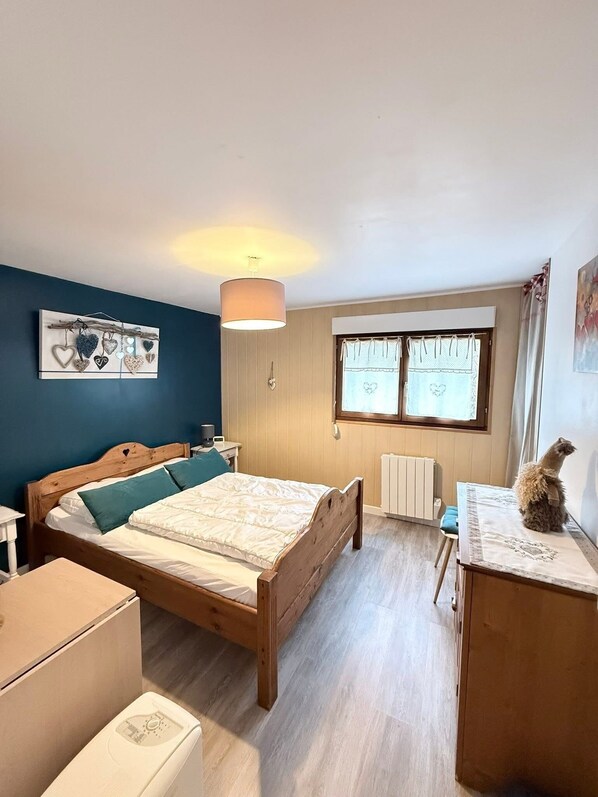 A bedroom with a wooden bed, a desk, a chair, a radiator, and a window with curtains. - Charming 3-room apartment in the hills above Gérardmer (Gérardmer)