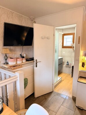A compact living space with a television mounted on the wall, a white door leading to a bathroom, and a small table with a lamp and decorative items. - Charming 3-room apartment in the hills above Gérardmer (Gérardmer)