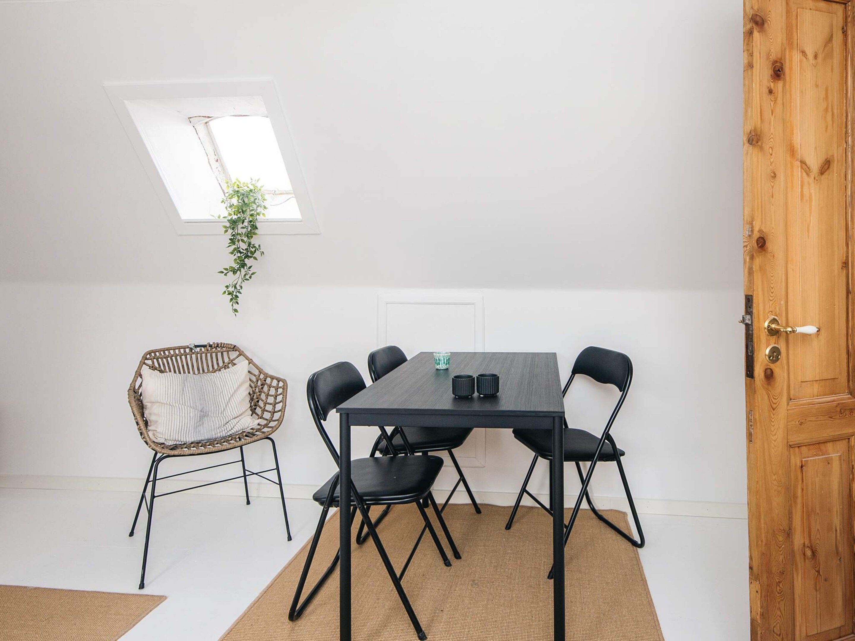 A room with a dining table, six chairs, a wicker chair, a small plant, a window, and a wooden door.