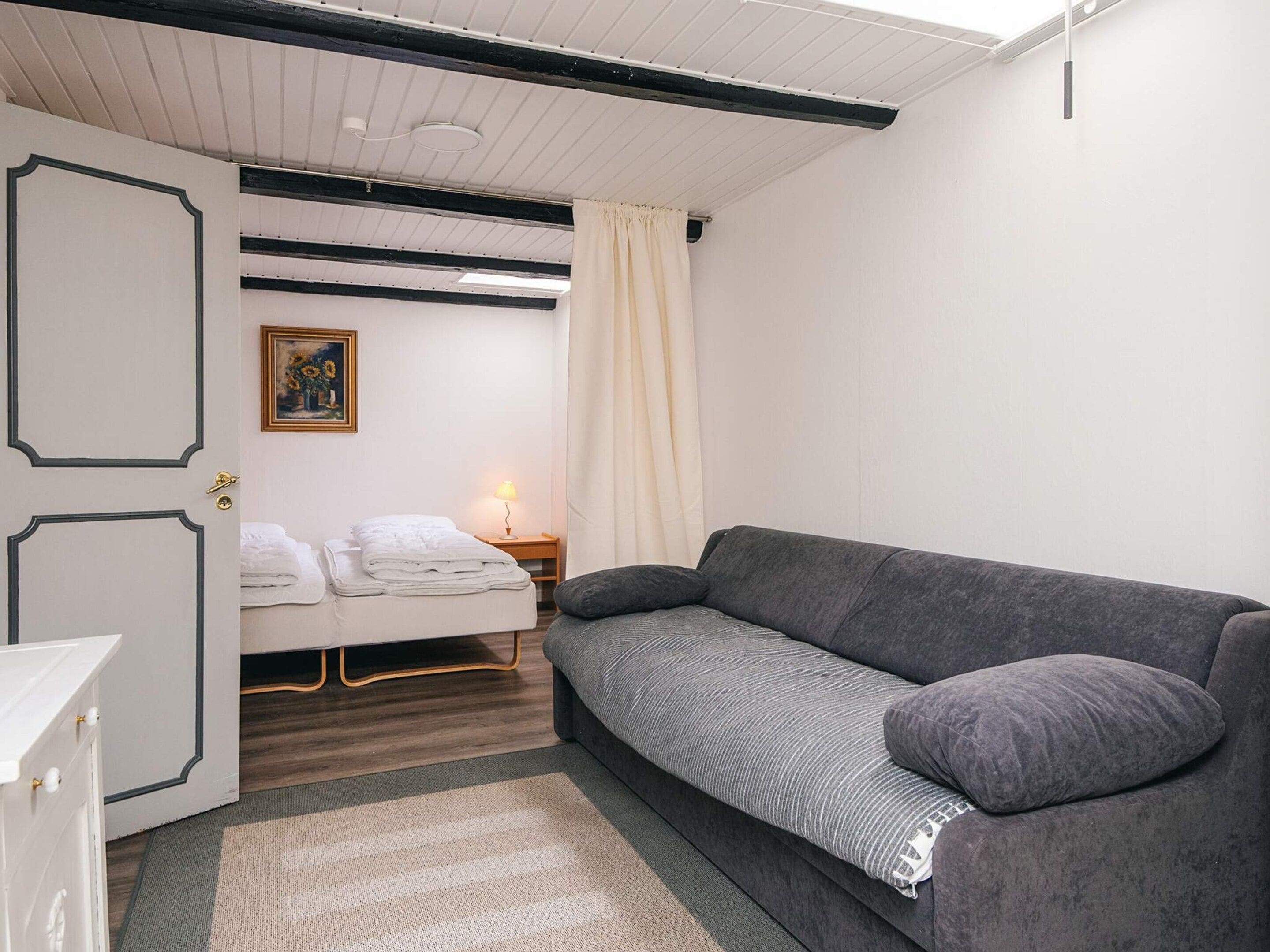 A room with a bed, a grey sofa, a white door, a painting, and a nightstand with a lamp.