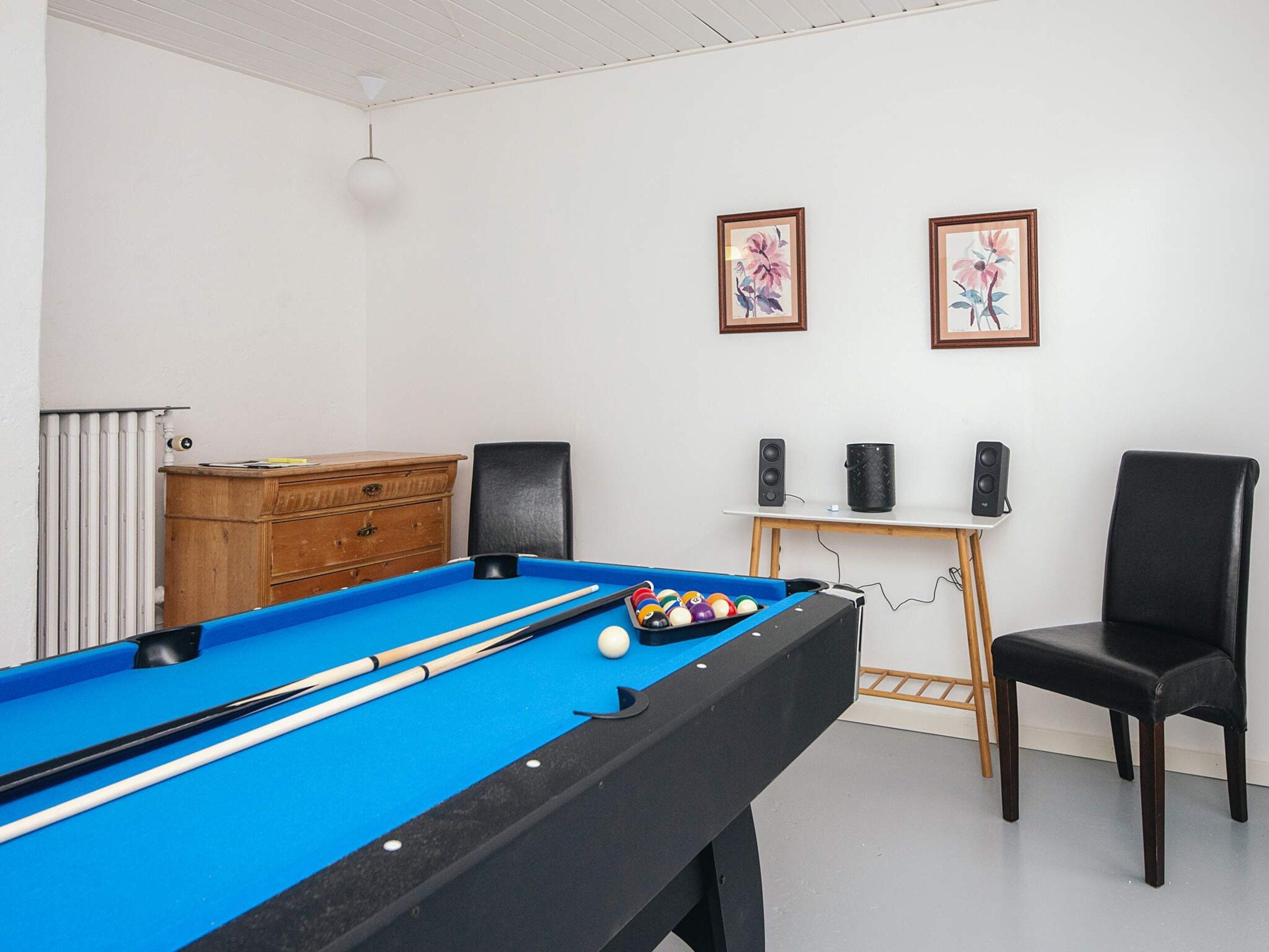 A pool table with balls and cues, a wooden dresser, a black chair, two framed floral paintings, and a speaker stand.