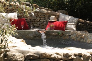 Eine Steinsäule mit einem kleinen Wasserfall, roten Kissen und einem Holztisch mit Glas und Krug.