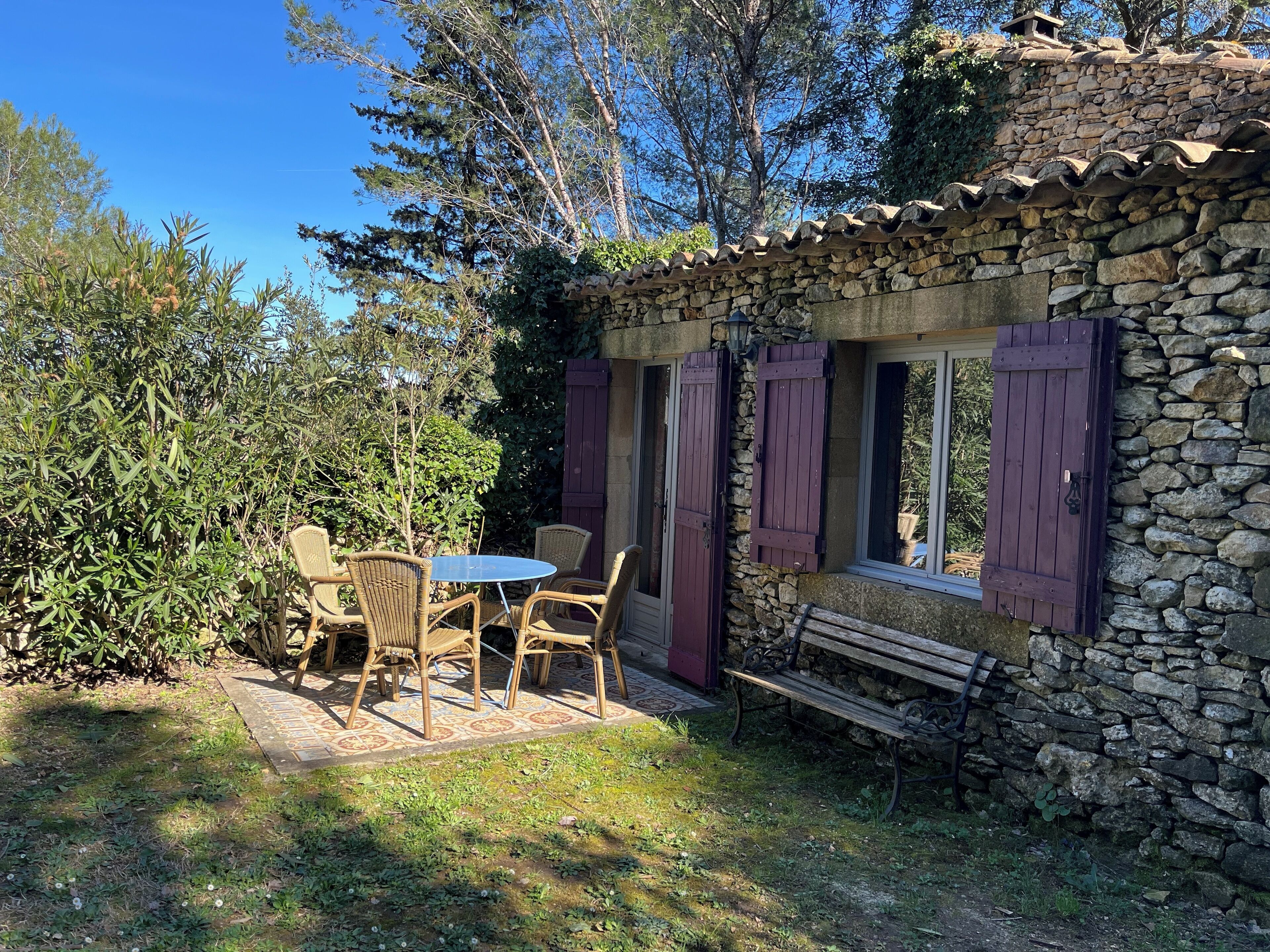 Un bâtiment en pierre doté d’un petit patio comprenant une table et des chaises, un banc et une fenêtre avec des volets.