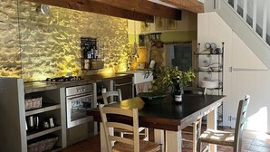A rustic kitchen with a wooden table and chairs, a stone wall backsplash, and a staircase with a white railing. - Mazet Carcarie, vacation home Provence, secluded location, stone house, nature, garden, dog (Montaren-et-Saint-Médiers)