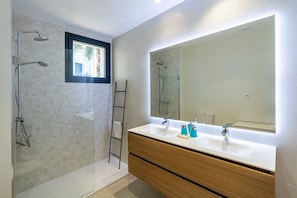 A modern bathroom with a glass-enclosed shower, a wooden vanity with a large mirror, and a window above the sink. - 6-guest Apartment Near Golf Course (Estepona)