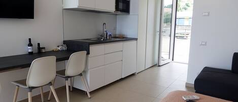 A compact kitchen with a modern design, featuring a built-in oven, a sink, and a microwave. There are two chairs and a small table with a remote control on it.