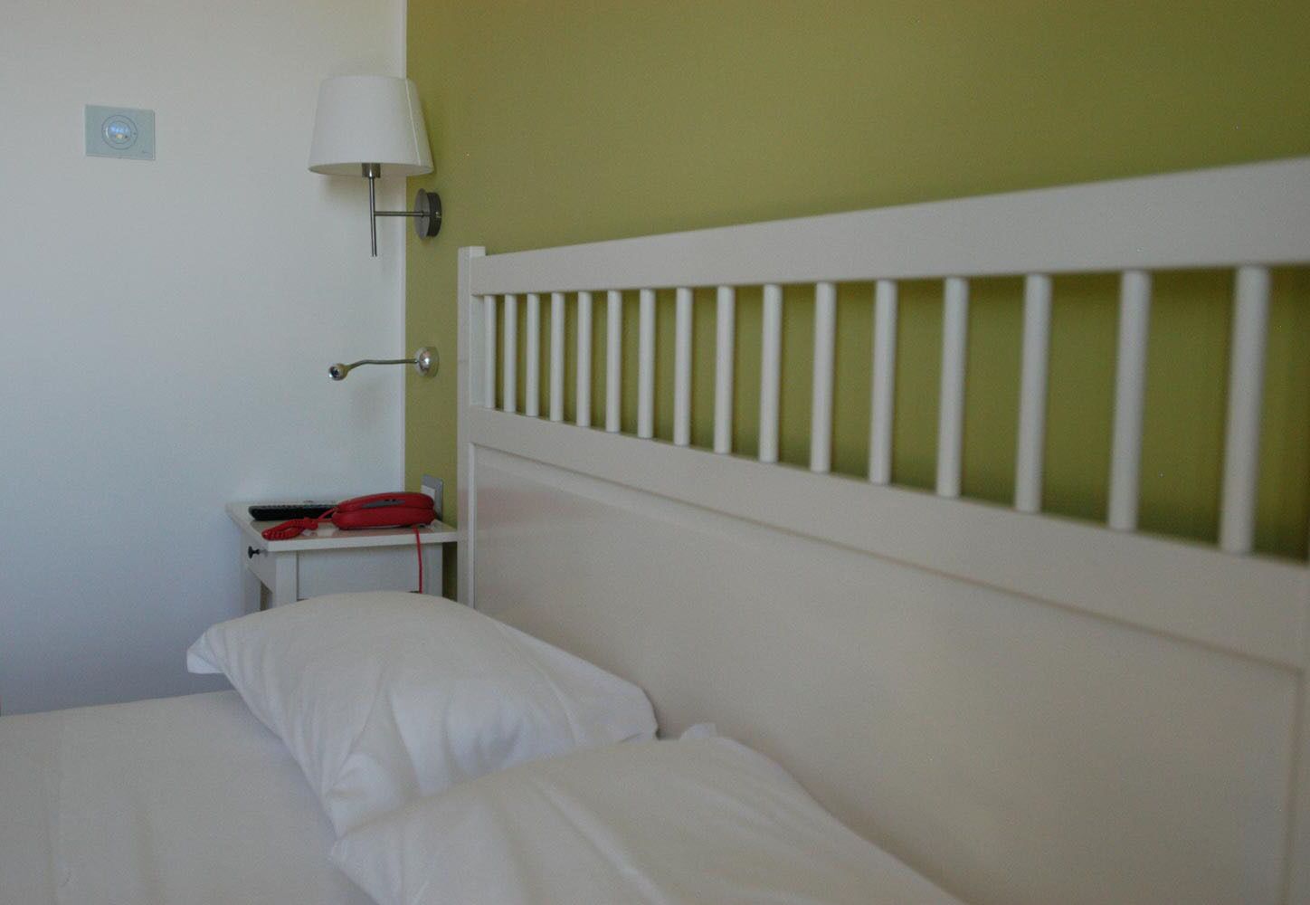 A bed with white bedding, a nightstand with a red object, a wall-mounted lamp, and a white headboard.