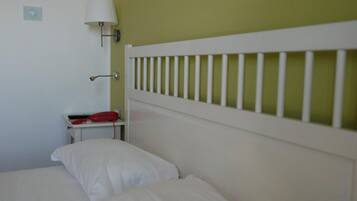 A bed with white bedding, a nightstand with a red object, a wall-mounted lamp, and a white headboard.