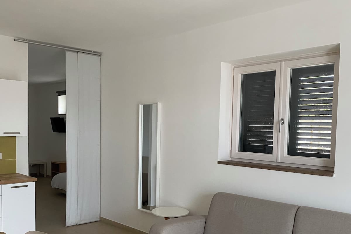 A modern living room with a sofa, a dining table with chairs, a kitchen area, and a window with blinds.