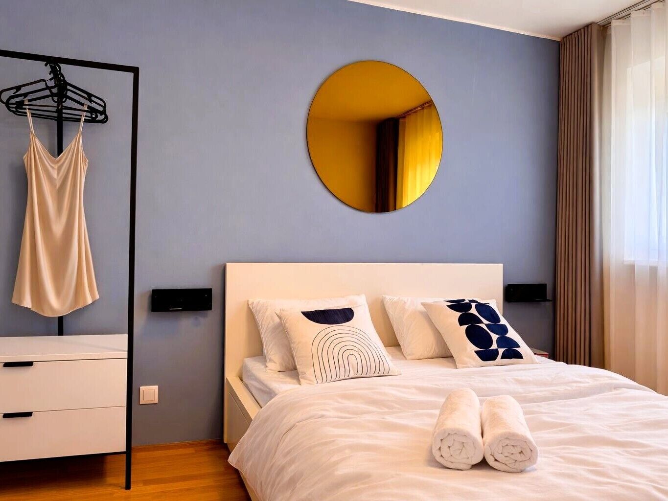 A modern hotel room with a bed, a circular mirror, a hanging garment rack, and a bedside table.