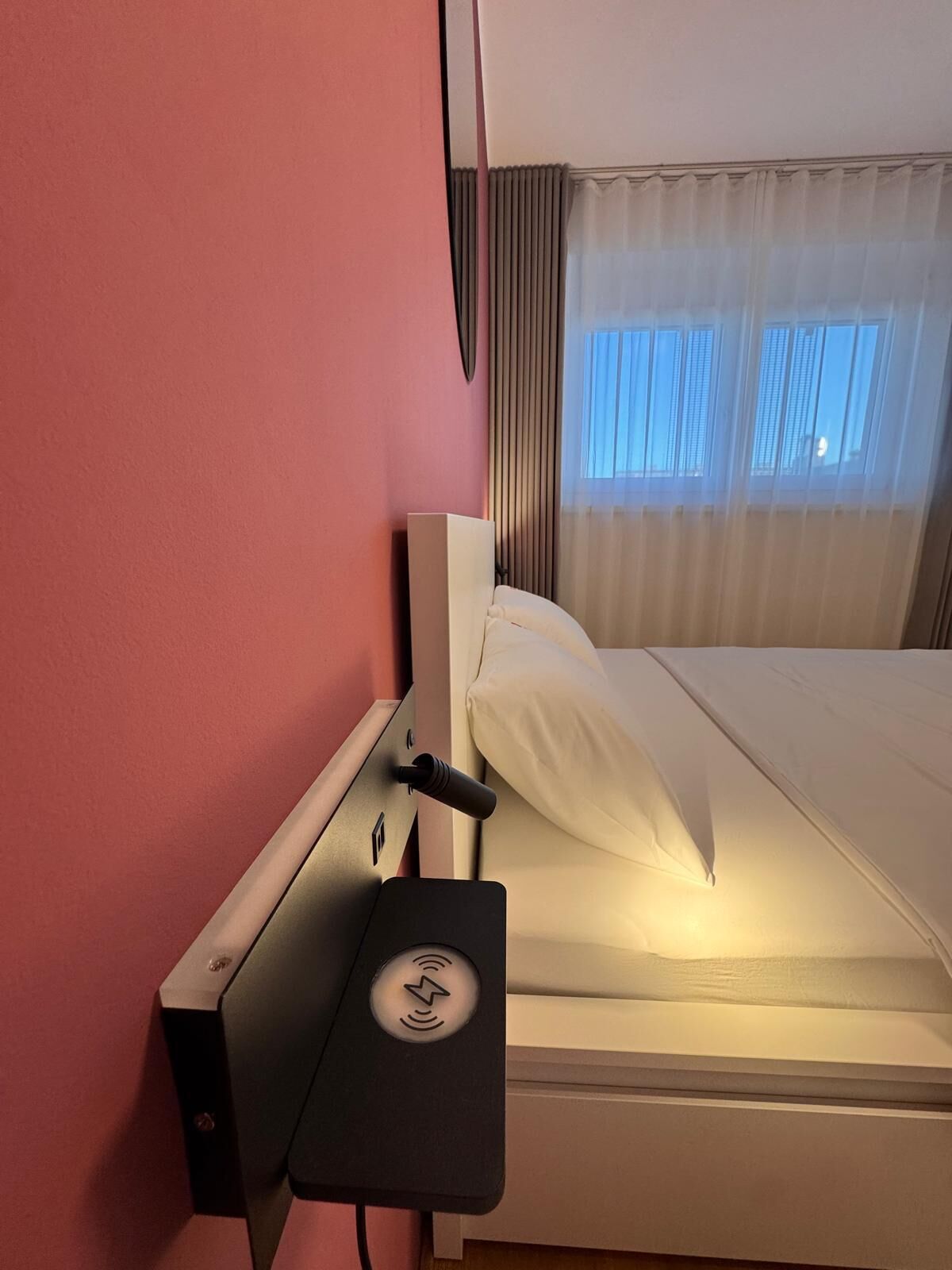 Una habitación de hotel con cama, mesita de noche con toma de corriente y ventana con cortinas.