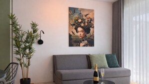 Een moderne woonkamer met een grijze bank, een ronde salontafel met een fles en glas, een groot kunstwerk aan de muur en een bamboe plant.
