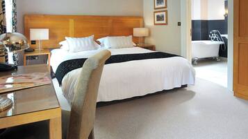 A hotel room with a large bed, a wooden headboard, two bedside tables with lamps, a desk with a chair, and a bathroom visible through an open door.