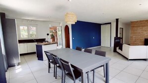 Une cuisine moderne avec des meubles blancs, un coin repas doté d’une grande table et de chaises, et un salon avec un canapé et une télévision.