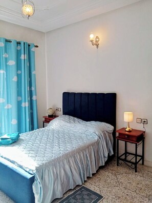 A bedroom with a bed, a nightstand, a lamp, a wall sconce, and a window with curtains.