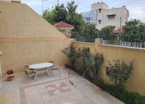 A patio with a table and chairs, a tiled floor, and a small garden area with bushes and a fence.