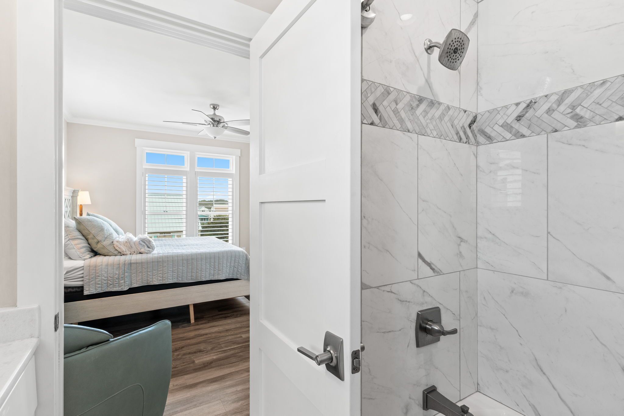 Ein Schlafzimmer mit Bett, einem Fenster mit Jalousien, einem Deckenventilator und einem Badezimmer mit Dusche und gefliesten Wänden.