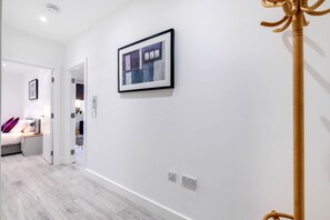 A hallway with a framed abstract painting, a coat rack, and a bed with pillows in the background. - Luxurious 1 Bed Serviced Apt. 02 with Wi-Fi (Corby)