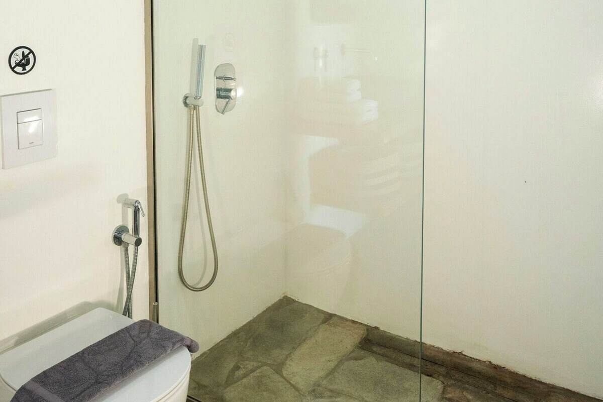 Une salle de bain avec une douche à parois de verre, des toilettes avec une serviette à portée de main et un interrupteur visible.