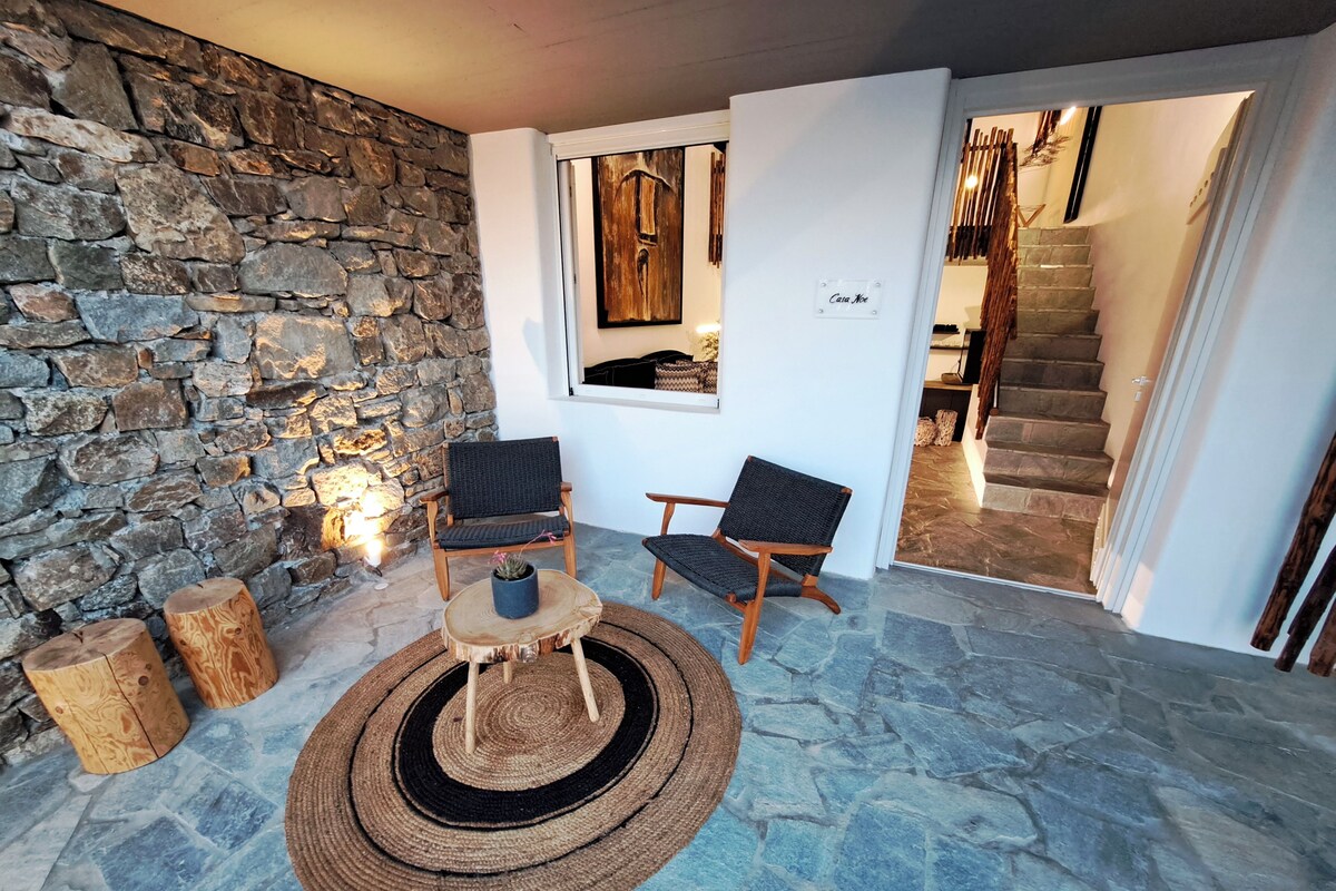 Un mur de pierres, deux fauteuils en osier, un tapis tissé et un escalier menant à un niveau supérieur.