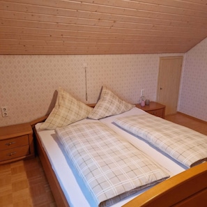 Ein Schlafzimmer mit Holzbalkendecke, zwei Betten mit Karo-Bettwäsche, einem Nachttisch, einer Kommode und einer Tür.