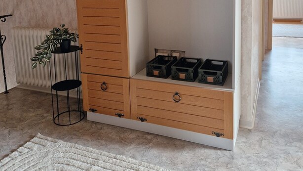 Ein Holzkleiderschrank mit mehreren Schubladen und einer Kleiderstange, platziert in einem Raum mit Heizkörper und einer kleinen Pflanze.
