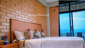 A neatly made bed with a wooden headboard, a nightstand with a vase, and a view of the mountains through a balcony. - Rocksana Fortress Yoga & Retreat (Mawanella)