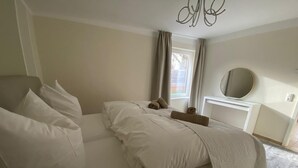 Ein Schlafzimmer mit Bett, Kissen, Nachttisch, Spiegel und einem Fenster mit Vorhängen.
