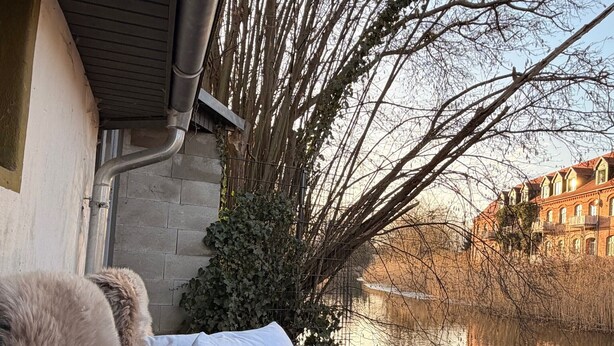 Eine Bank mit einem Fellüberwurf und einem Handtuch, mit Blick auf einen ruhigen Fluss, im Hintergrund ein Gebäude.