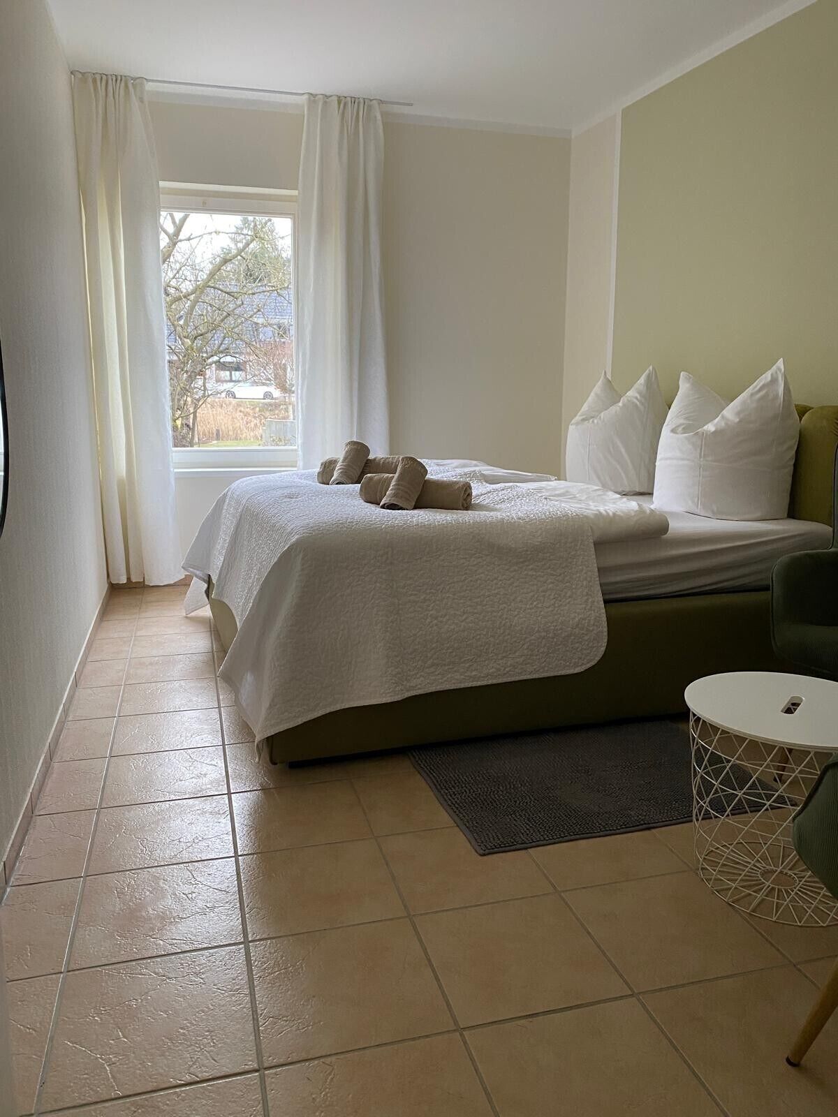 A hotel room with a bed, a small round table, a chair, and a window with curtains.