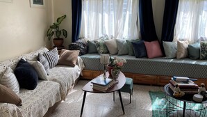 Un salón acogedor con un sofá estampado, una mesita de centro con libros, una planta en maceta y una ventana con cortinas traslúcidas.