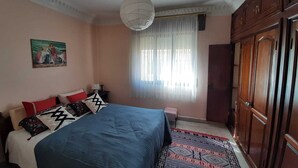 Un dormitorio con cama, mesita de noche, lámpara, ventana con cortinas, un cuadro en la pared y armario.