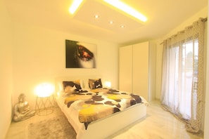 A bedroom with a bed, bedside lamps, a Buddha statue, a painting, and a window with curtains. - Beachside Apartment With Jacuzzi & Pool (Marbella)