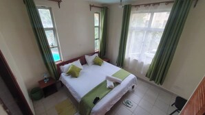 Een net opgemaakt bed met groen-wit beddengoed, een klein nachtkastje met een lamp, en uitzicht op het groen buiten door een raam met groene gordijnen.