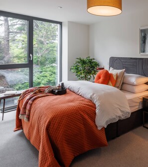 A modern bedroom with a large window, a bed with orange bedding, a bedside table, and a potted plant. - The Ironworks (Dublin)