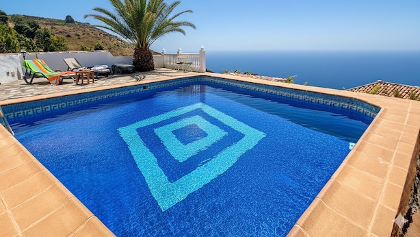 Ein Schwimmbecken mit geometrischem Muster, umgeben von Liegestühlen und einer Palme, mit Blick auf eine Küstenlandschaft.