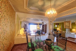 Une pièce avec un papier peint à motifs, un lustre, des fauteuils à coussins verts, un grand vase décoratif et une vue sur une autre pièce où se trouvent un canapé et des lampes.
