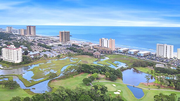 Een golfbaan met waterhindernissen, omgeven door woongebouwen en een strand.
