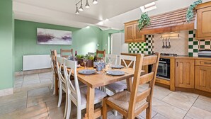 A dining area with a wooden table set for six, surrounded by white chairs. A radiator is visible against a green wall, and a kitchen with wooden cabinets and a checkered backsplash is in the background. - Tackley House - pet friendly near Oxford (Near Oxford)