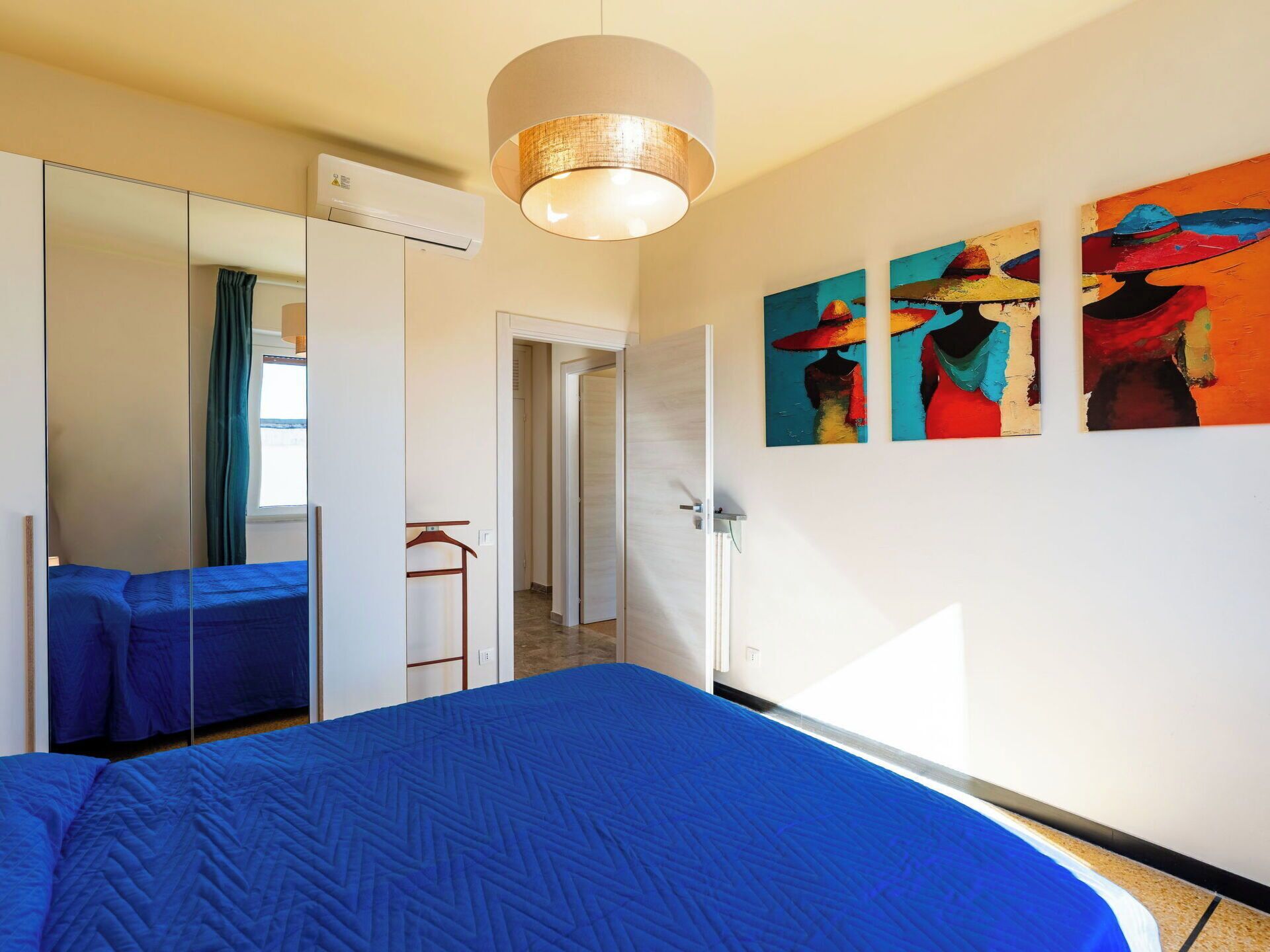 Une chambre d’hôtel avec un grand lit, une armoire, un miroir et trois tableaux colorés accrochés au mur.