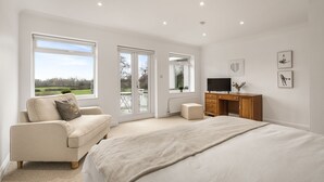 A spacious bedroom with a large bed, a sofa, a TV, and a desk. There are large windows and a view of the outdoors.