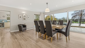 A modern dining area with a wooden table and black chairs, large windows with a view of trees, and a kitchen in the background.
