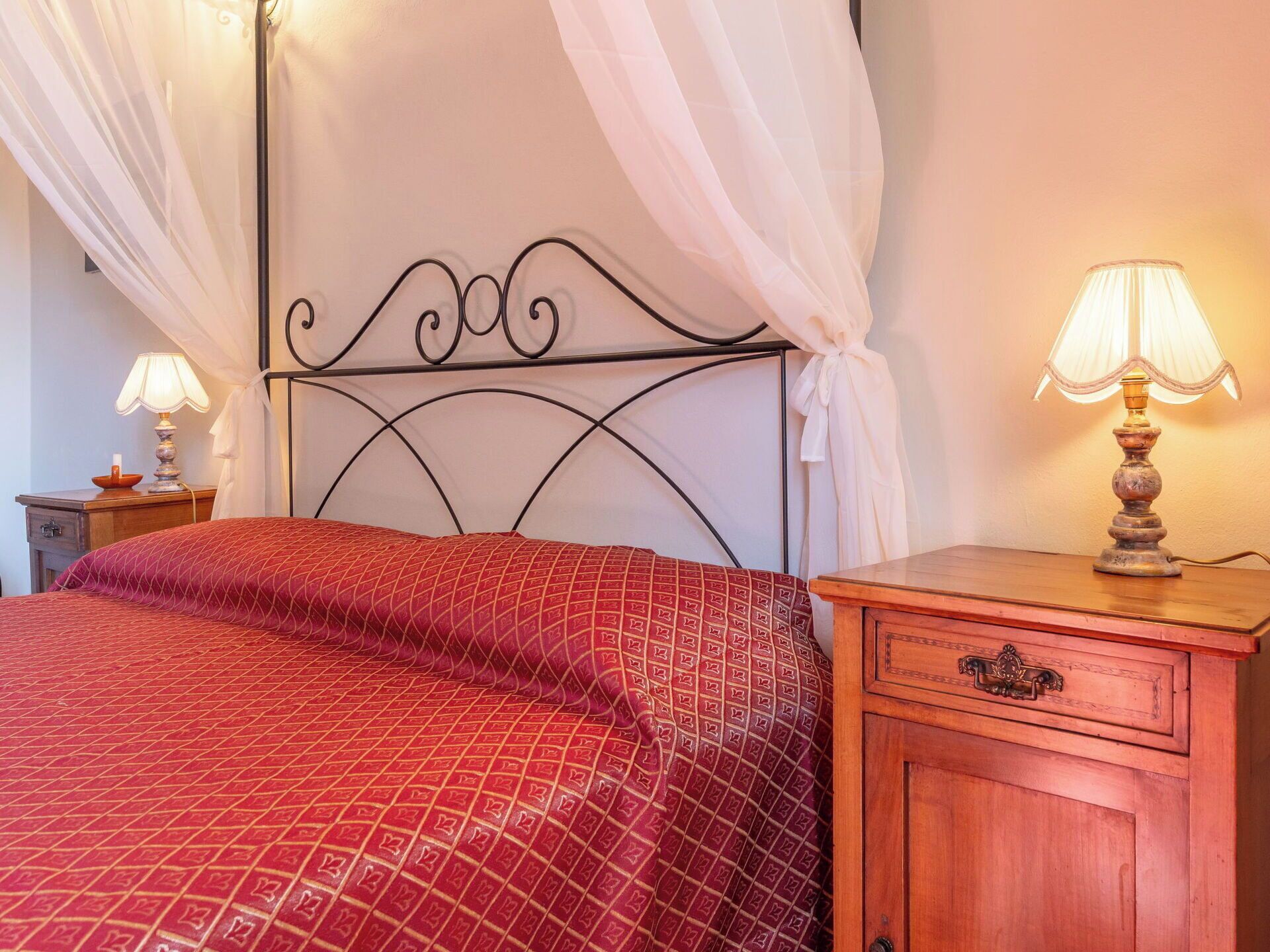 Un lit recouvert d’une couvre-lit à motifs rouges, une table de chevet en bois avec une lampe et une commode en bois avec un tiroir.