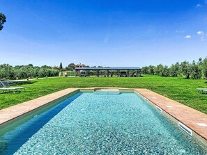 Ein rechteckiger Pool mit einer gefliesten Terrasse, umgeben von einer Rasenfläche und einem überdachten Pavillon.