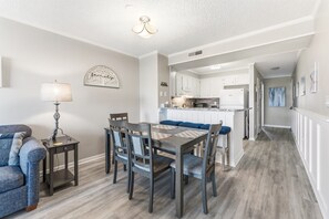 A modern kitchen with white cabinets, a dining area with a wooden table and chairs, and a hallway leading to a living room. - Pelicans Landing 213 3 BR Beach/Pool (Myrtle Beach)