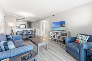 A living room with a sofa, a coffee table, a TV, and a dining area with chairs and a table. - Pelicans Landing 213 3 BR Beach/Pool (Myrtle Beach)