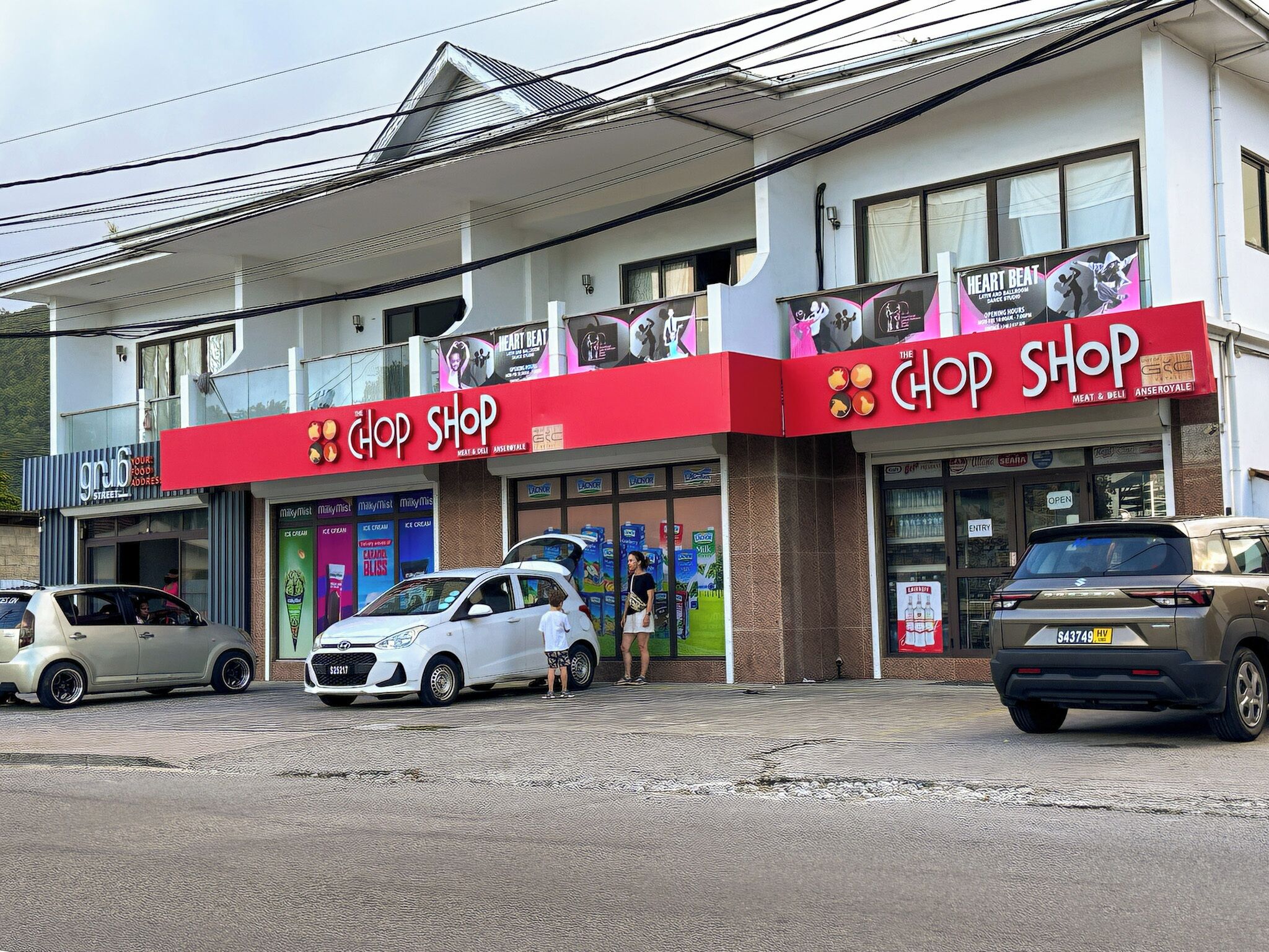 Ein Gebäude mit roter und weißer Fassade, zwei Geschäfte mit den Schildern 'The Chop Shop' und 'Heart Beat' sowie parkende Autos davor.