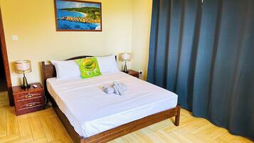 Una cama bien hecha con un almohadón, una mesita de noche con lámpara, un cuadro colgado en la pared y una ventana con cortinas.