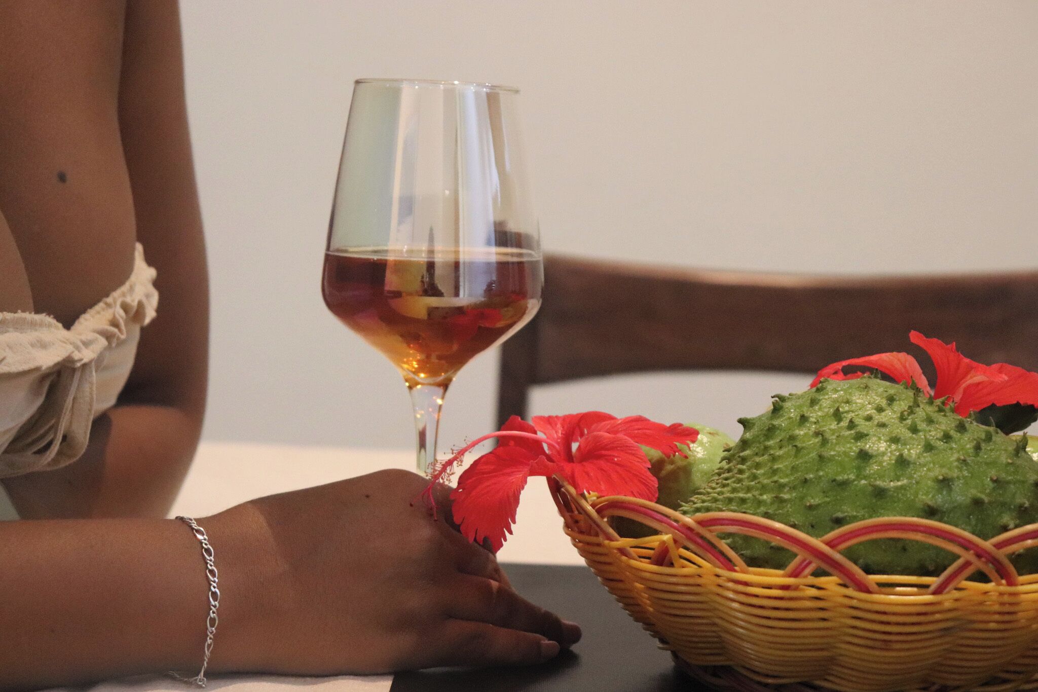 Una persona sentada a una mesa con una copa de vino tinto, una cesta de mimbre con frutas y una silla.